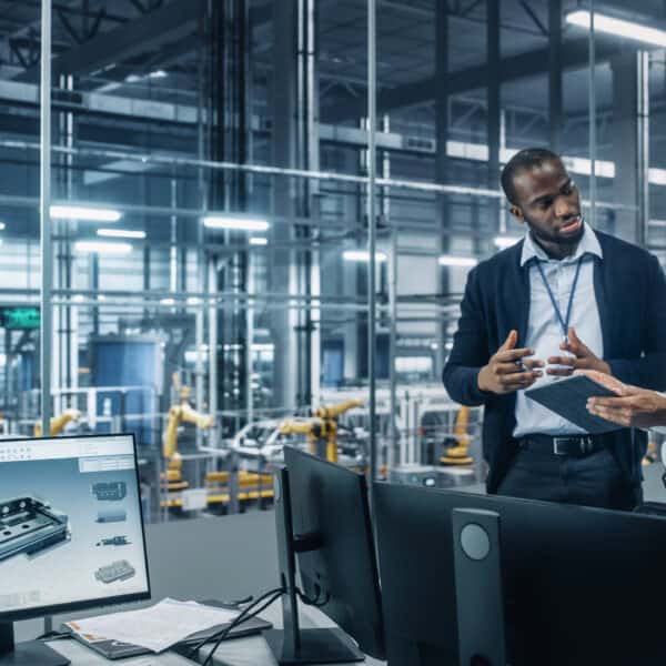 Two Diverse Automotive Industrial Engineers Talking About Vehicle Production while Standing in Office at a Car Assembly Plant. Industrial Specialists Discuss Work Projects on Tablet Computer.