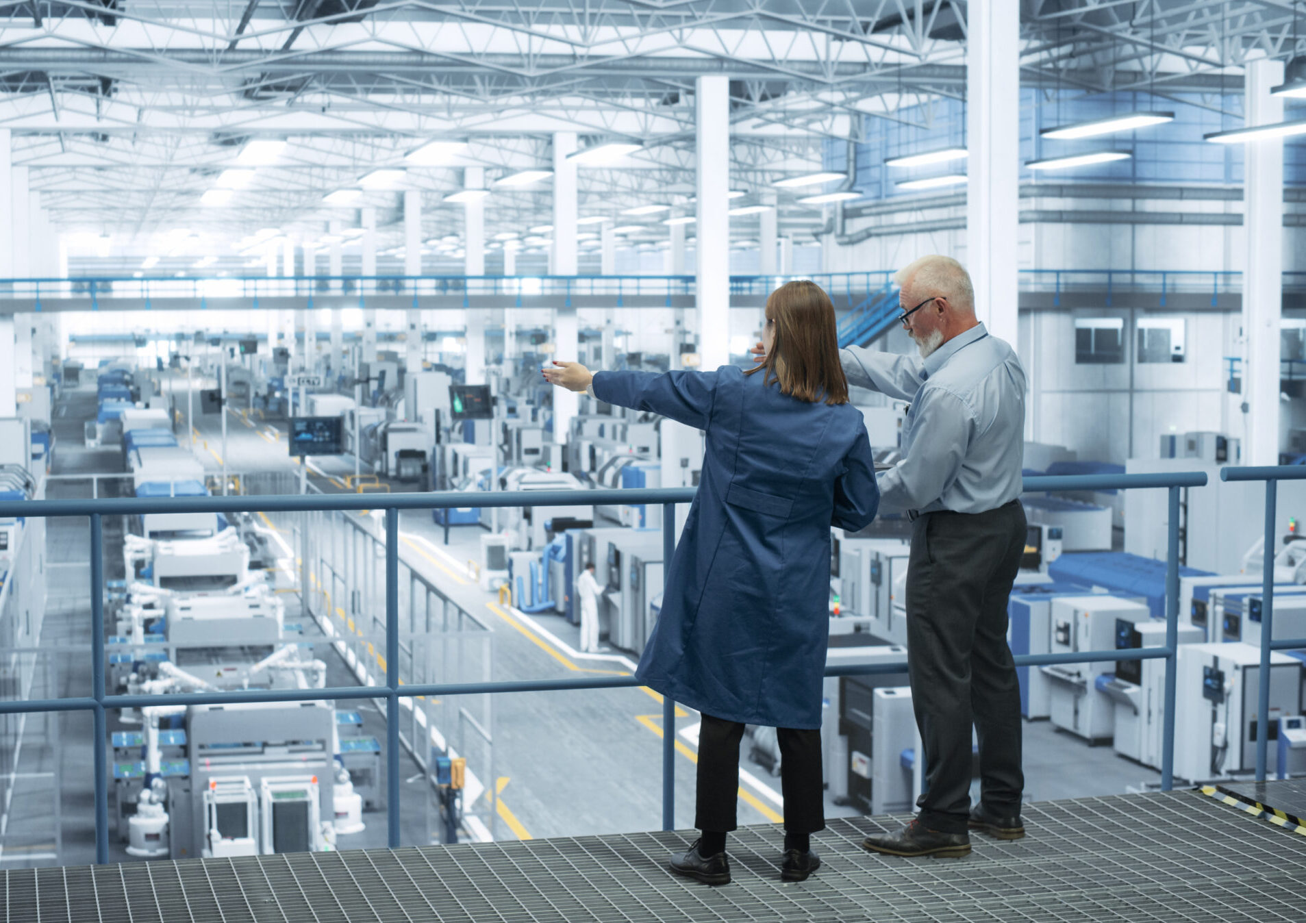Experienced Male and Female Engineers Standing on a Platform with Their Back to Camera, Using Laptop Computer and Discussing Production at a Modern AI Automated Electronics Manufacture